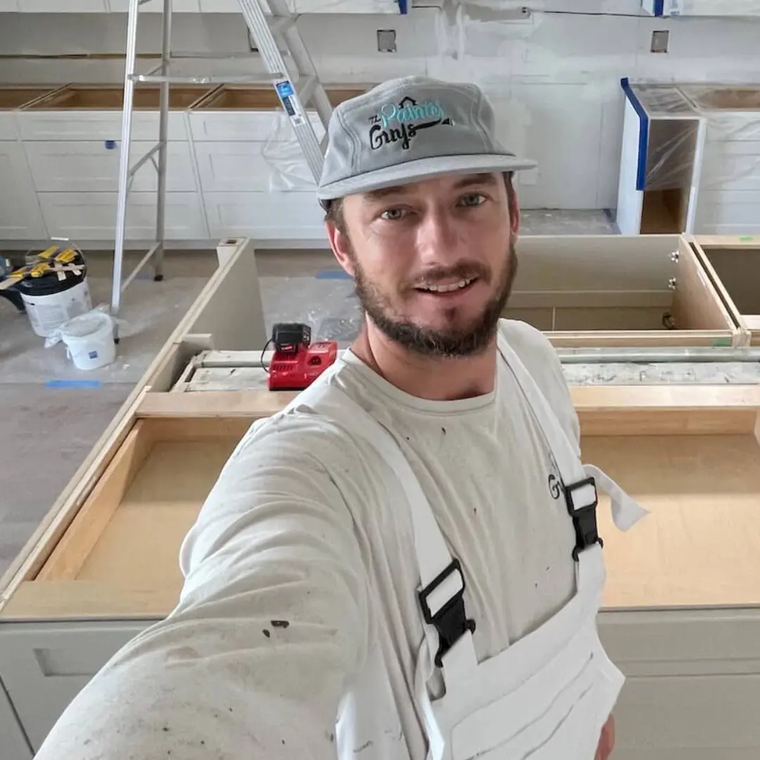 Painter Guys owner smiling on a cabinet painting job site wearing a Painter Guys cap and painter overalls
