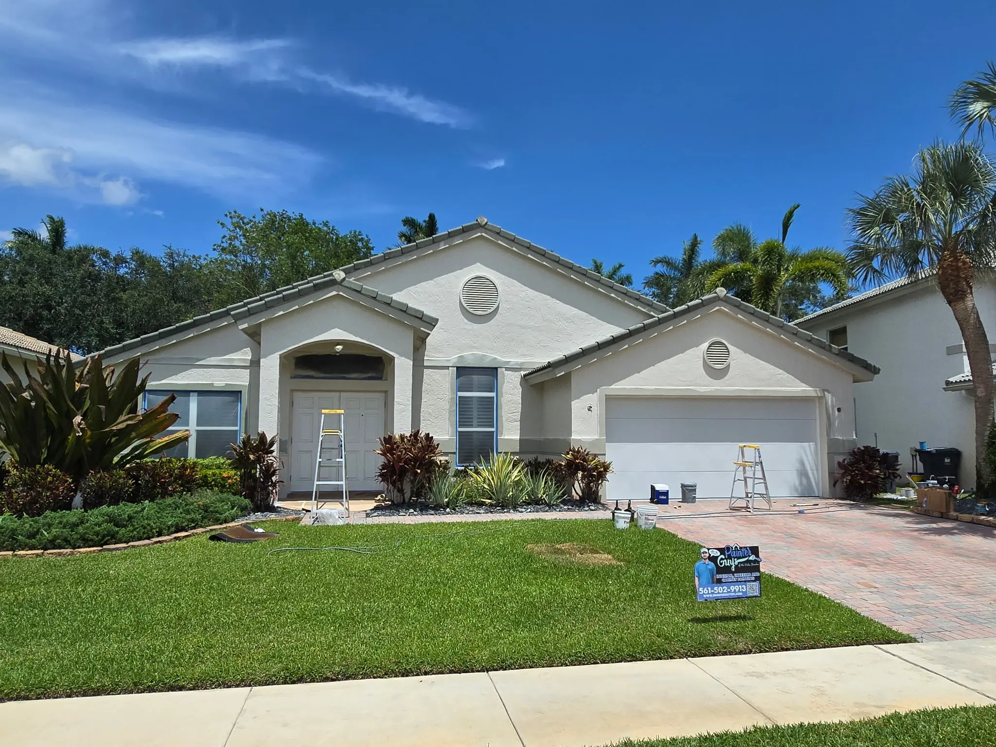 Painter Guys crew painting the exterior of a single-story stucco home with ladders and equipment on the brick paver driveway in Wellington FL