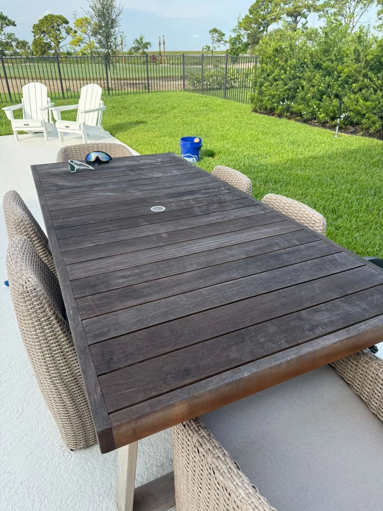 Weathered outdoor teak dining table with wicker chairs on a painted white concrete patio with manicured lawn in Wellington FL