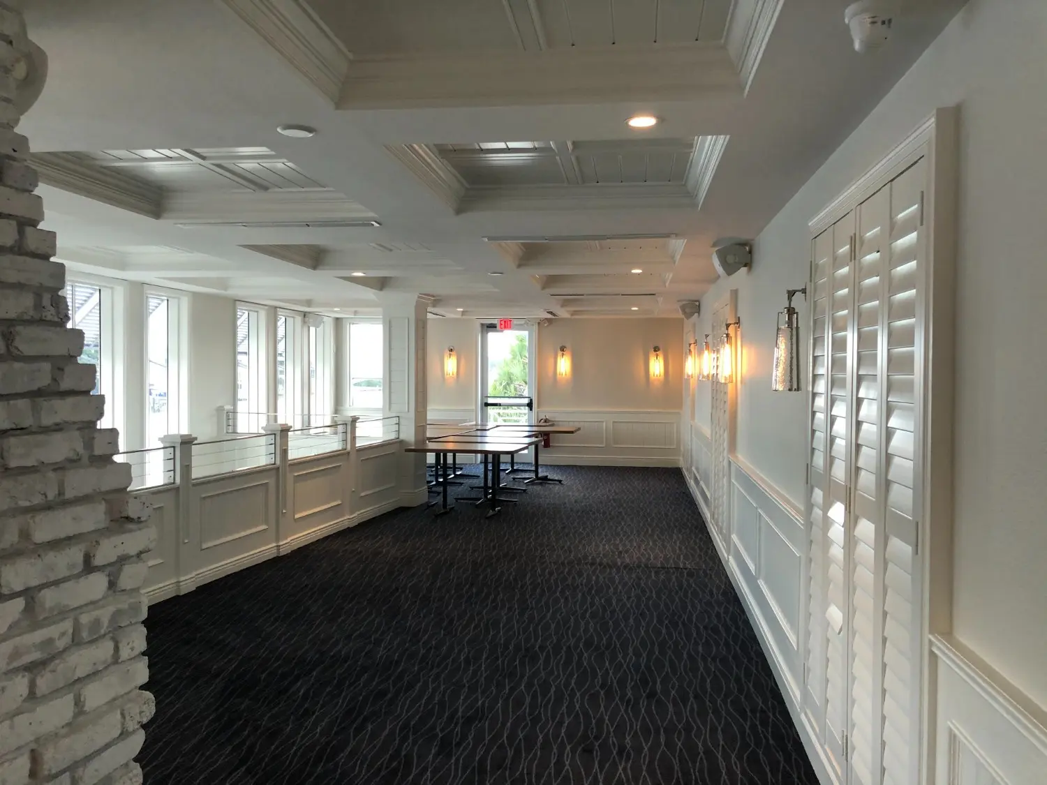 Commercial dining space with white coffered ceiling, wainscoting, wall sconces, plantation shutters and exposed brick column