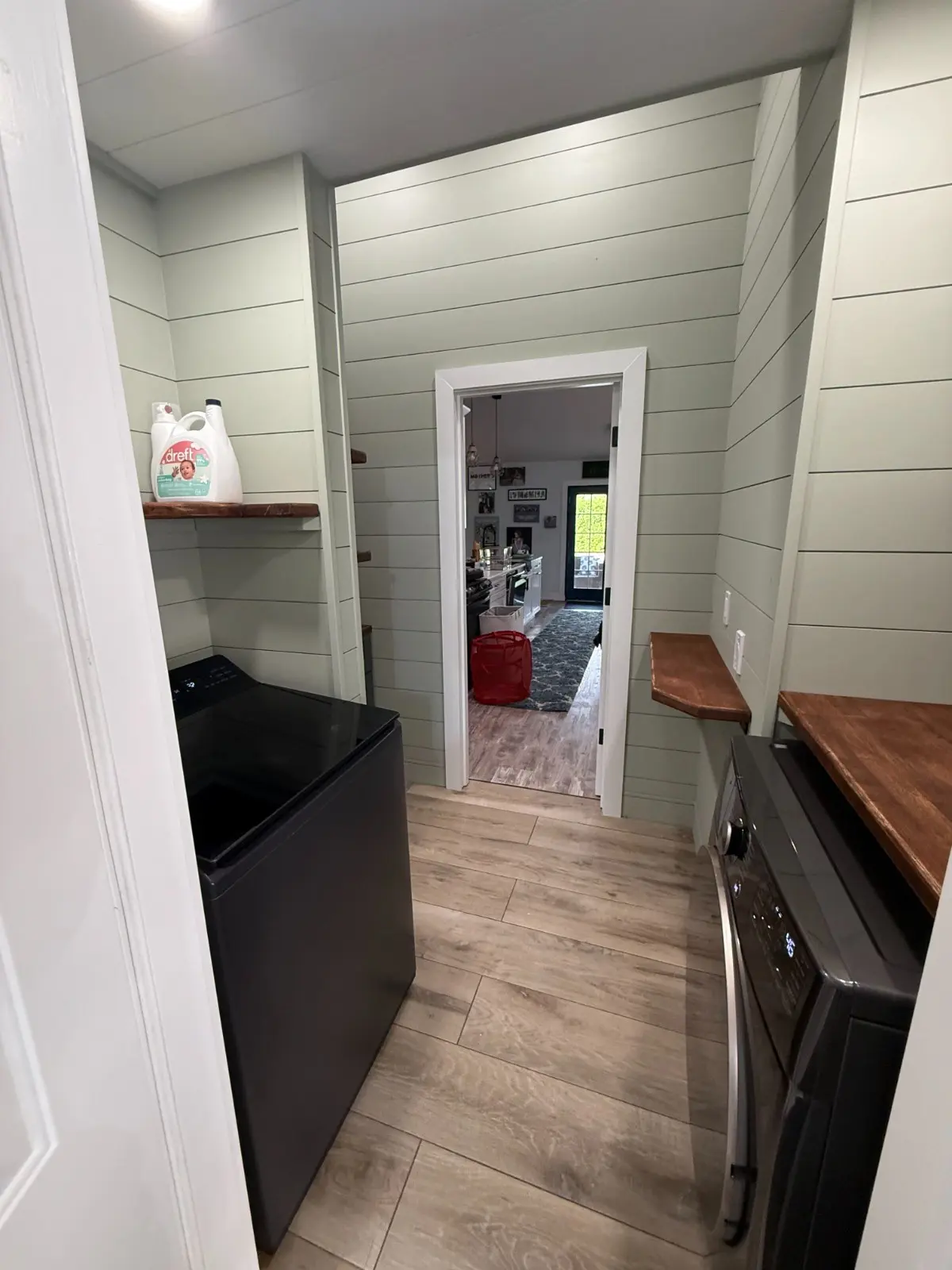 Sage green shiplap laundry room with washer and dryer, wood countertop shelving and open doorway to kitchen