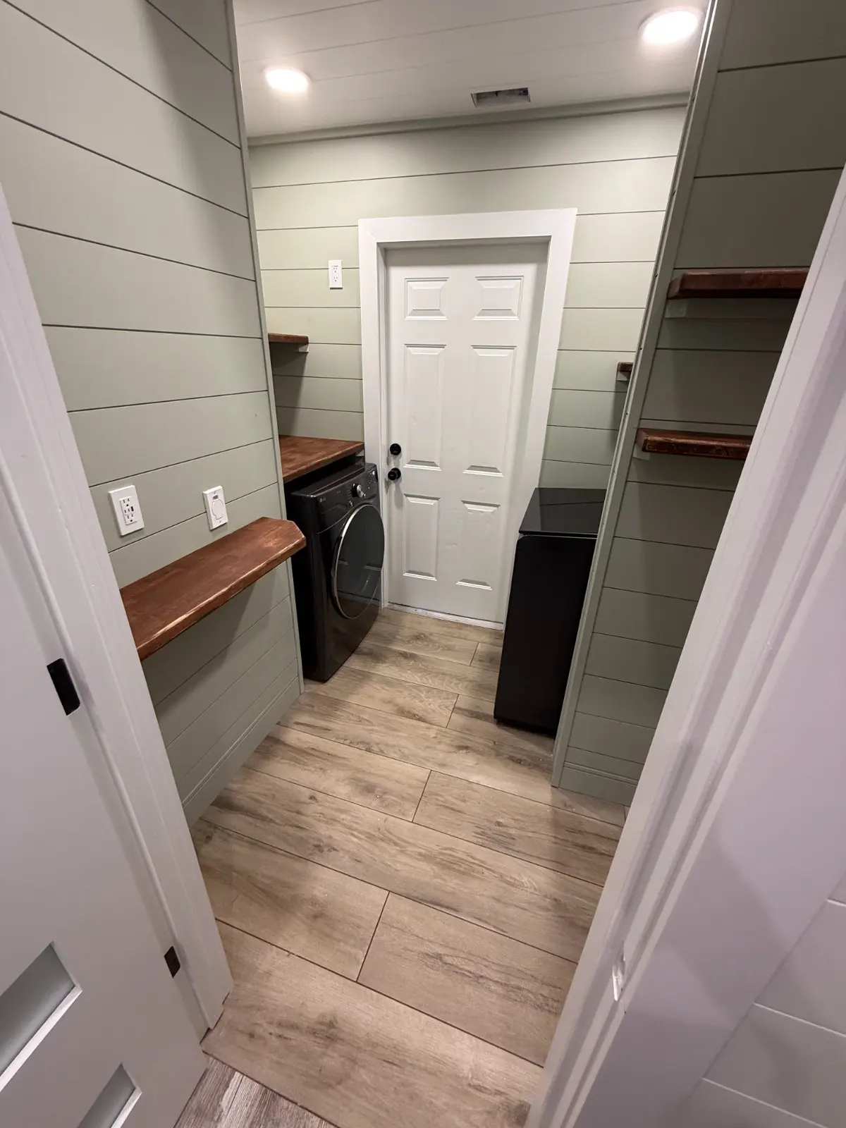 Sage green shiplap laundry room with wood countertop shelving, front-load washer and light wood tile floors