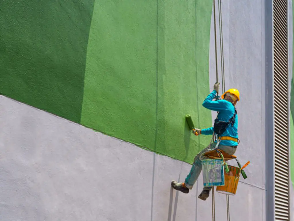 Commercial painter on rope access equipment painting the exterior of a large building.