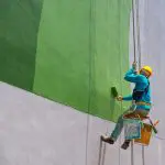 Commercial painter on rope access equipment painting the exterior of a large building.
